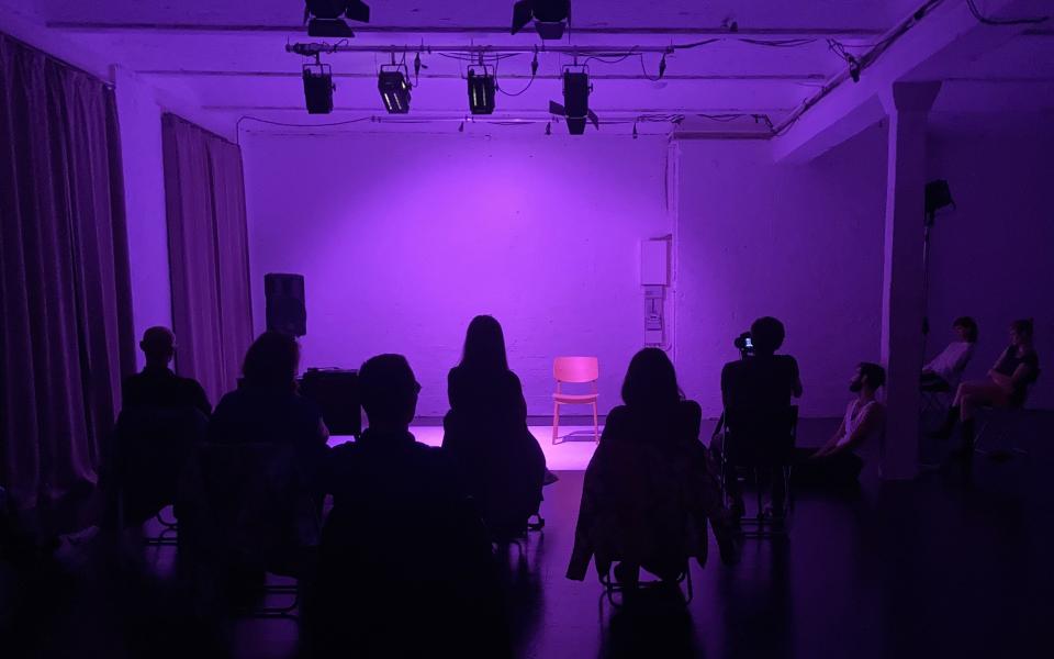 Aufnahme einer in Violett beleuchteten Bühne. Die Bühne, die hinter dem Publikum zu sehen ist, ist leer.