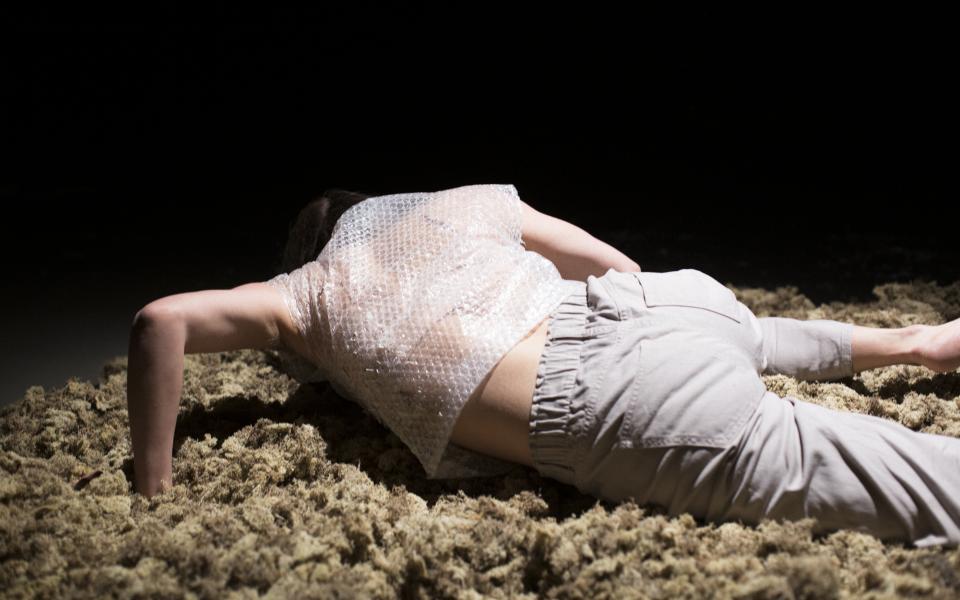 A person lies on a light surface of organic material - moss. The person is turned away from, props himself up a bit with his hands and keeps his head lowered. The person wears light pants and a short-sleeved shirt made of bubble wrap. The background is black.
