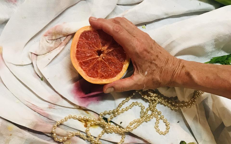© Julia Bosch Auf einem bekleckerten Tischtuch. Eine faltige Hand hält eine aufgeschnittene Blutorange. Um sie herum Essensreste und eine Goldkette.
