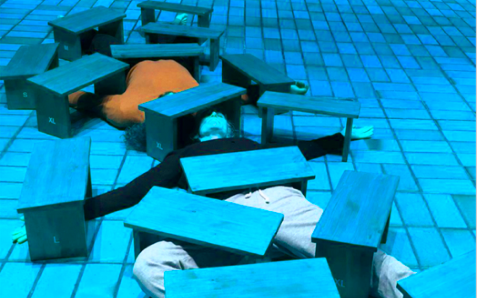 © ON URGENCY - Marcozzi Contemporary Theatre Zwei Körper liegen unter vielen Holzbänkchen, so dass nur ein Oberkörper in orange sowie Beine in beige erkennbar sind. Der Boden ist von Kacheln durchzogen und das Bild schimmert in einem blauton. s