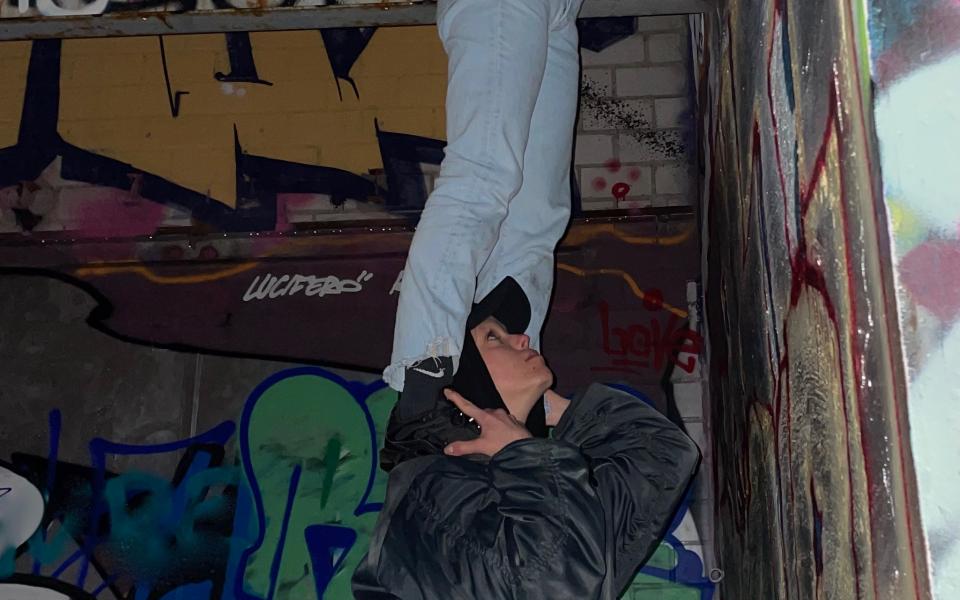 © Josephine Findeisen In front of a wall with graffiti, a person stands on the shoulders of another person and reaches up to the ceiling. The person below is also looking up.