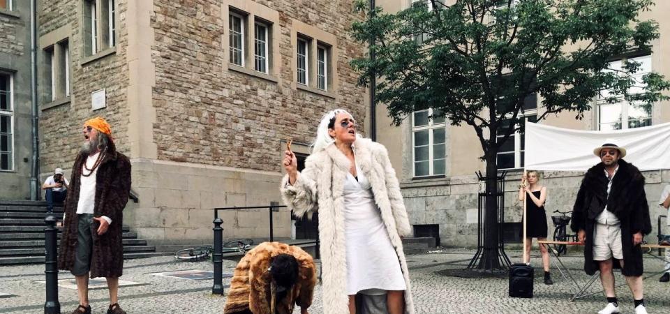 Vier Schauspieler in Pelzmänteln schreien auf einem Platz