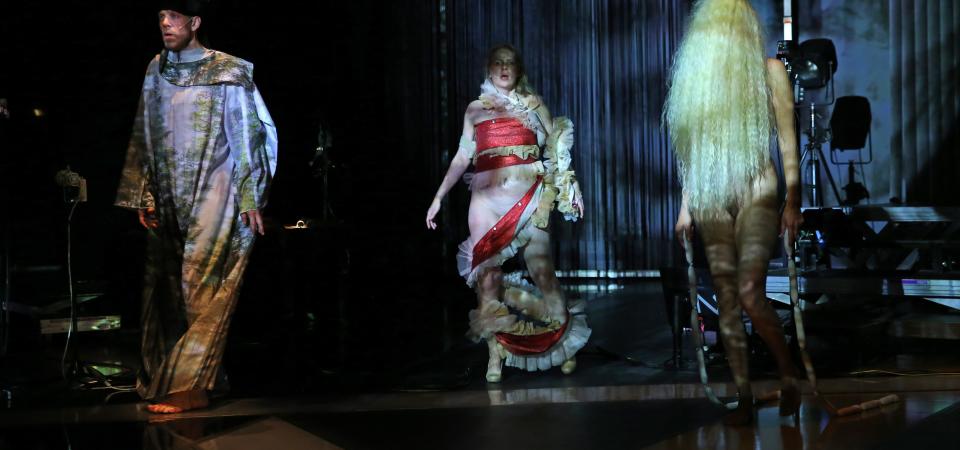 Zwei Performer:innen von She She Pop stehen in Kostümen auf der dunklen Bühne. Eine Performerin ist nackt, ihr Körper wird von einer blonden Langhaarperücke bedeckt. 
